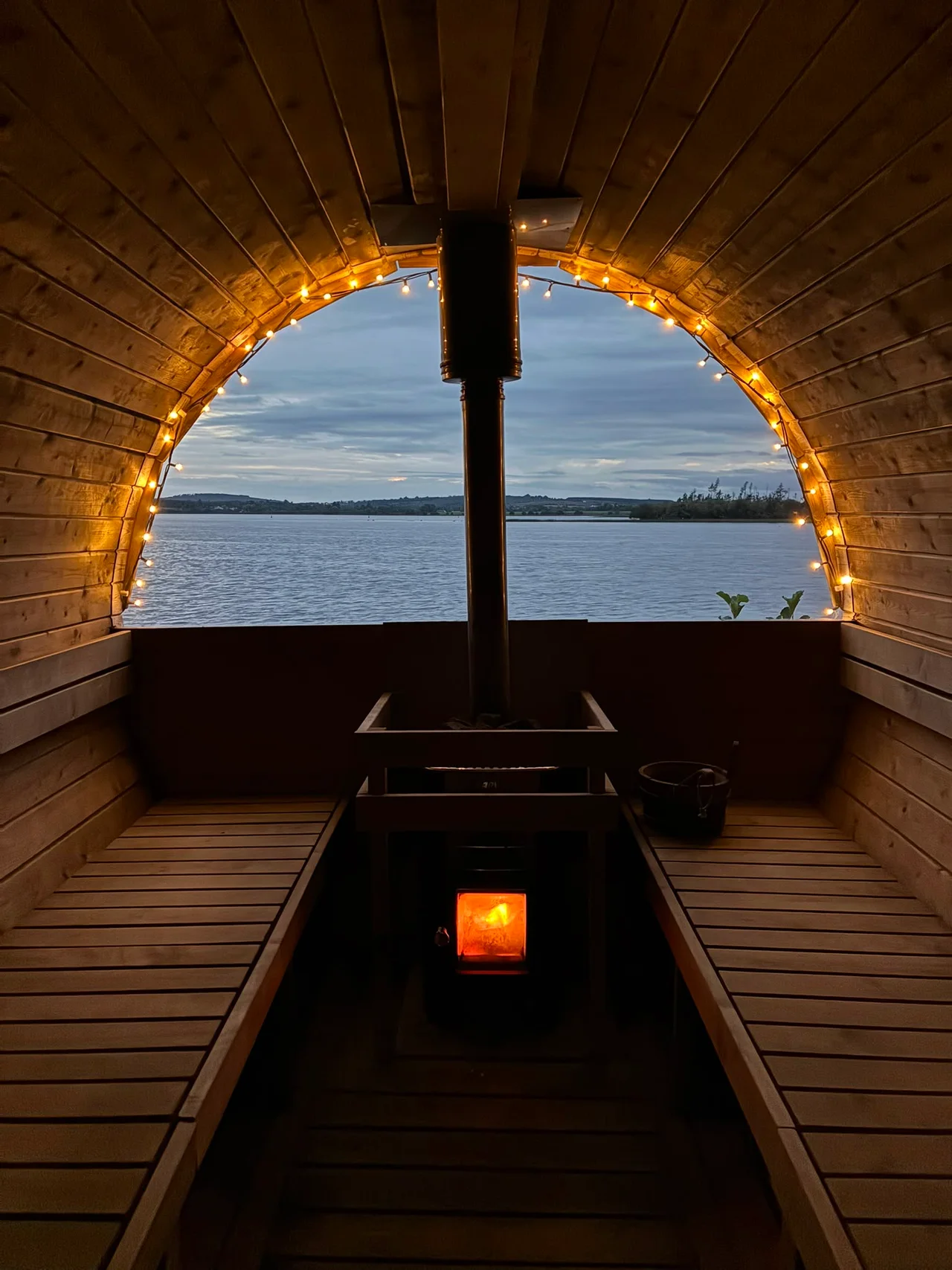 Fairy lights and a glowing fire frame a view of the lake from the sauna
