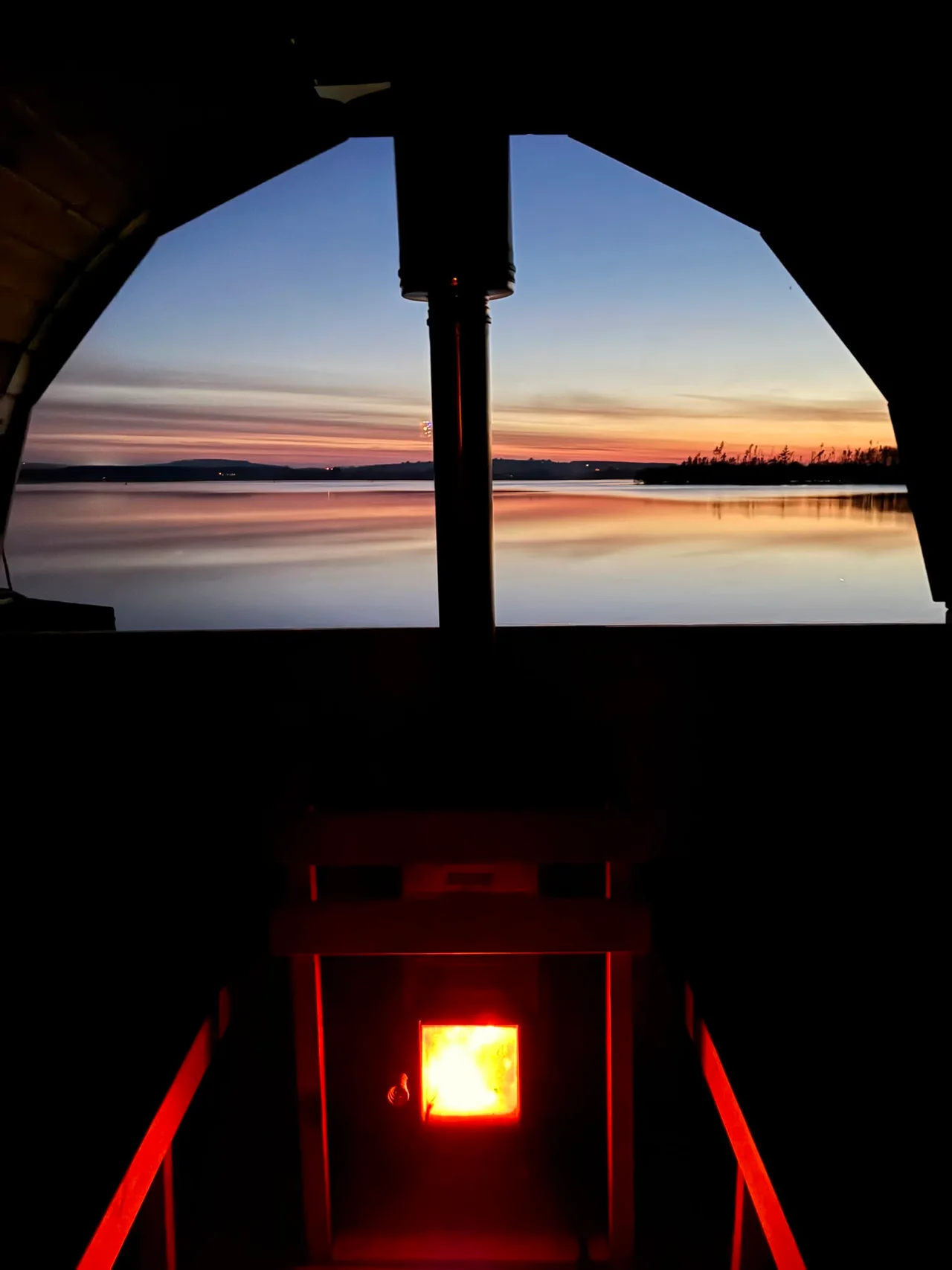 A glowing wood stove beneath the sauna window at dusk